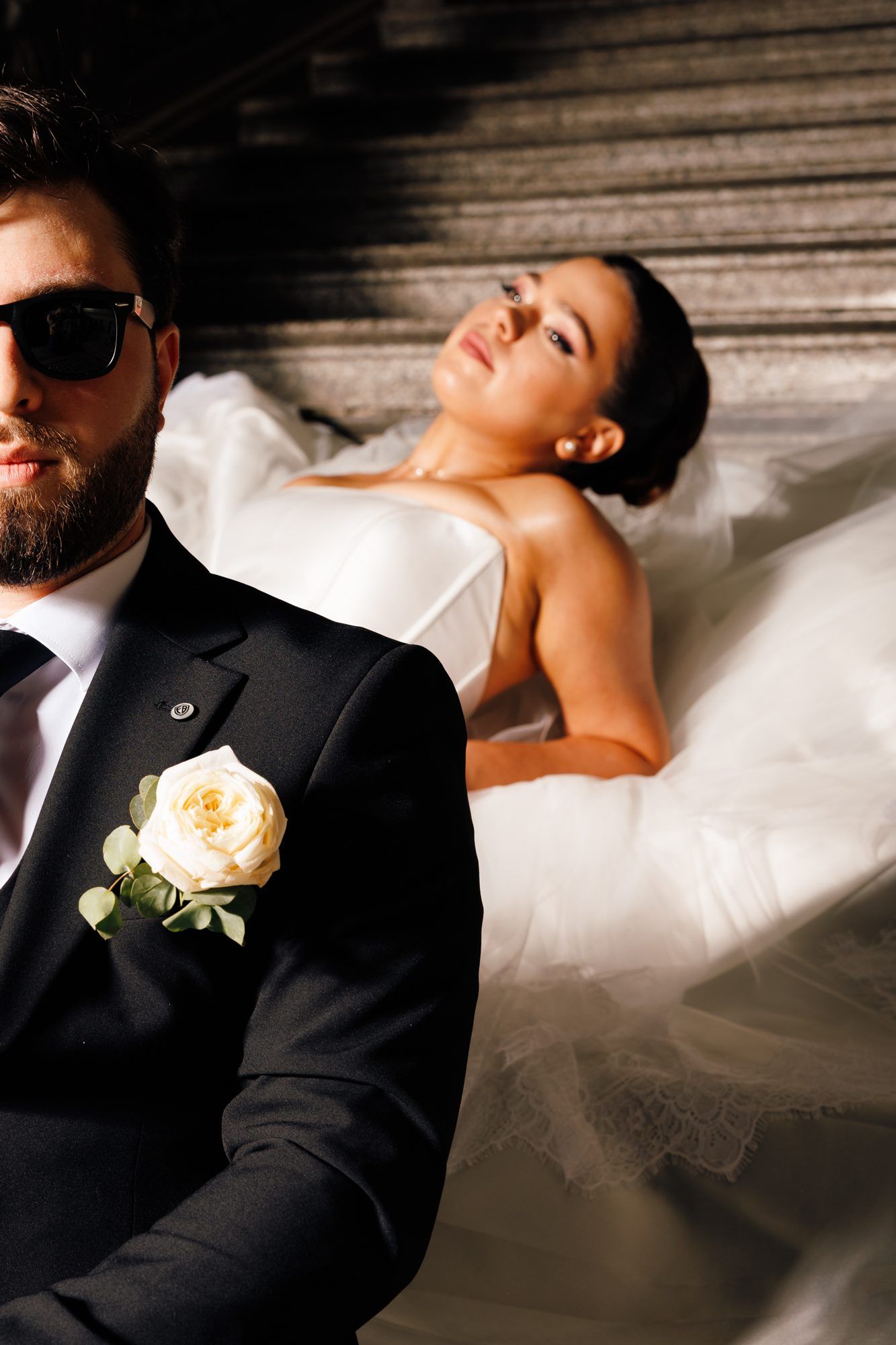 Groom in sunglasses with a white boutonnière, the bride reclining on the stairs behind him