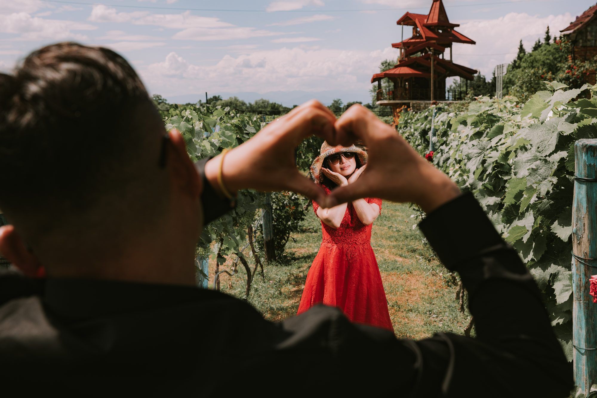 A man frames a woman in a red dress with his hands in a heart shape, in a vineyard